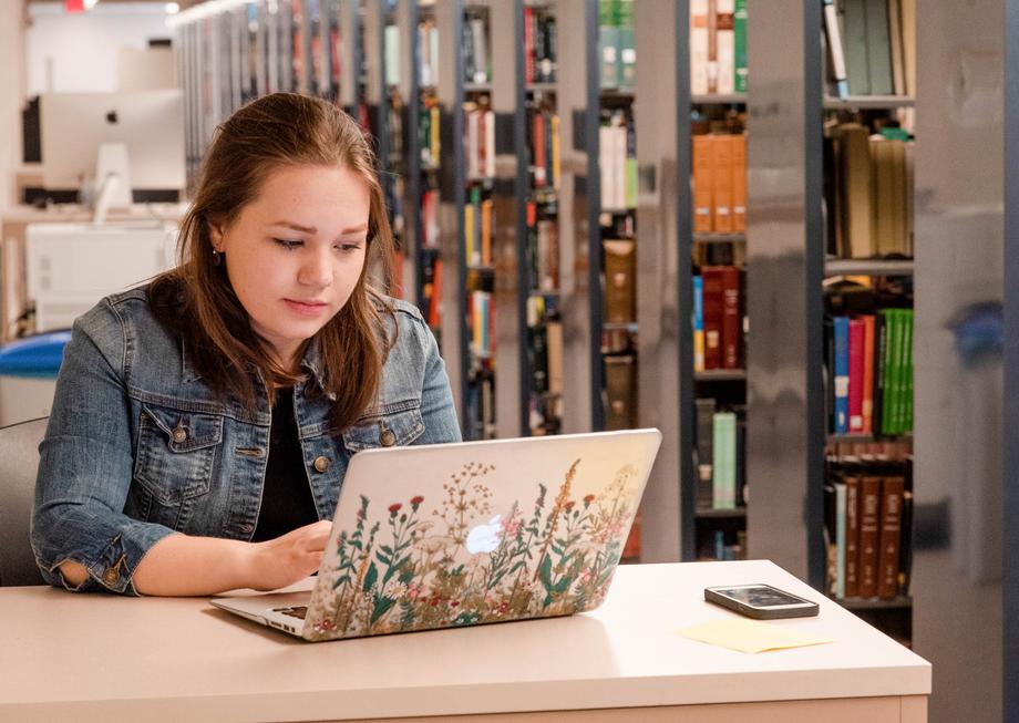 Student using her laptop at a table next to the book stacks.