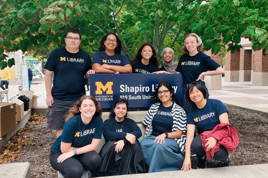 A group of 9 students in U-M Library t-shirts standing around the Shapiro Library sign