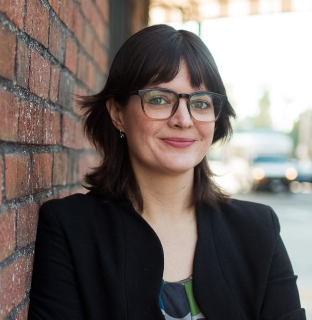 photo of smiling person with glasses and dark hair leaning against a brick wall