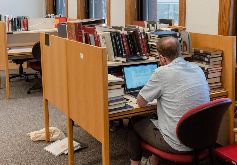 Student working on their laptop at an open carrel surrounded by stacks of books.