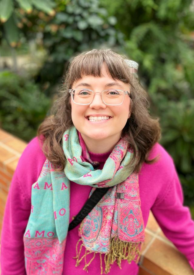 White woman with shoulder length brown hair, glasses wearing a pink sweater and colorful scarf. She is standing in front of a lush, plant background. 