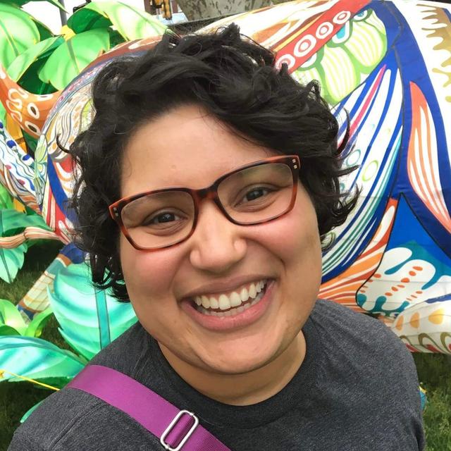 Photograph of Vitalina Robinson, a middle-aged latine woman with light brown skin, wearing glasses and smiling in front of a colorful background. 