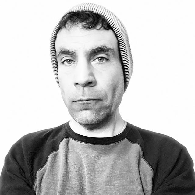 A black and white picture of Craig from the shoulders up.  Craig is wearing a long sleeved shirt and a winter hat, though it is unclear whether it is actually winter in the photo.