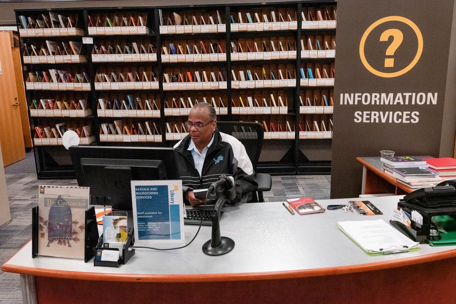 Smiling person sitting at a desk with a sign that says Information Services and stacks with current serials behind them