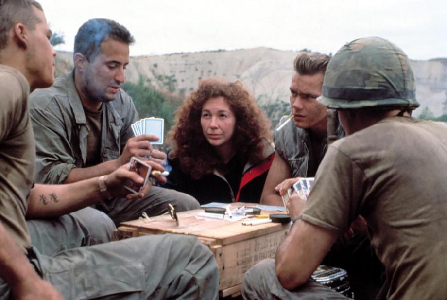 woman in the middle of four men sitting outside around a table playing cards